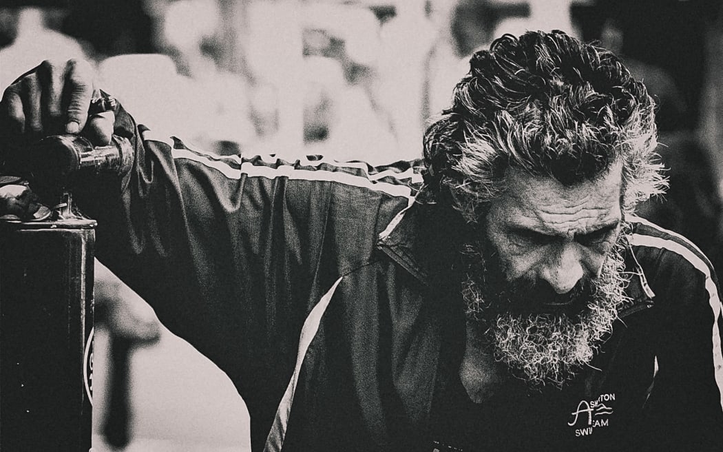 It is a black an white photo of a man (possibly homeless, but very stylish) leaning on a wall looking down towards the ground.