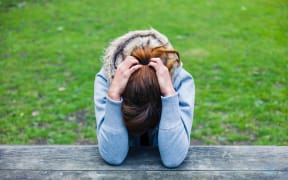 A young woman - sad or upset - sits on a bench with her head in her hands.