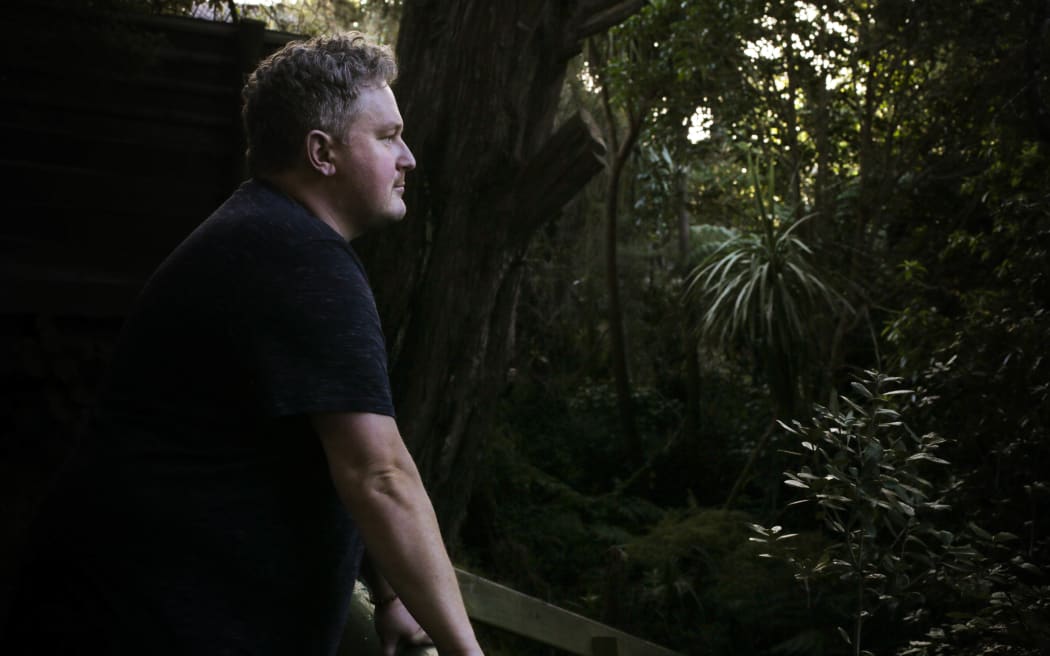 Lyall Carter, a tall man with curly blond hair, leans on a wooden railing overlooking dark green bush near his home in West Auckland.