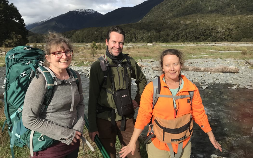 Three people in tramping gear and carrying backpacks stand in front of a gravel riverbed, with forest-cloaked mountains rising in the background to a cloudy grey sky.