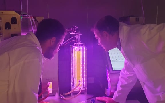 Two men in white lab coats look at a glass tube filled with something yellow-green. The room is bathed in purple light and other scientific machines are displays are visible in the background.