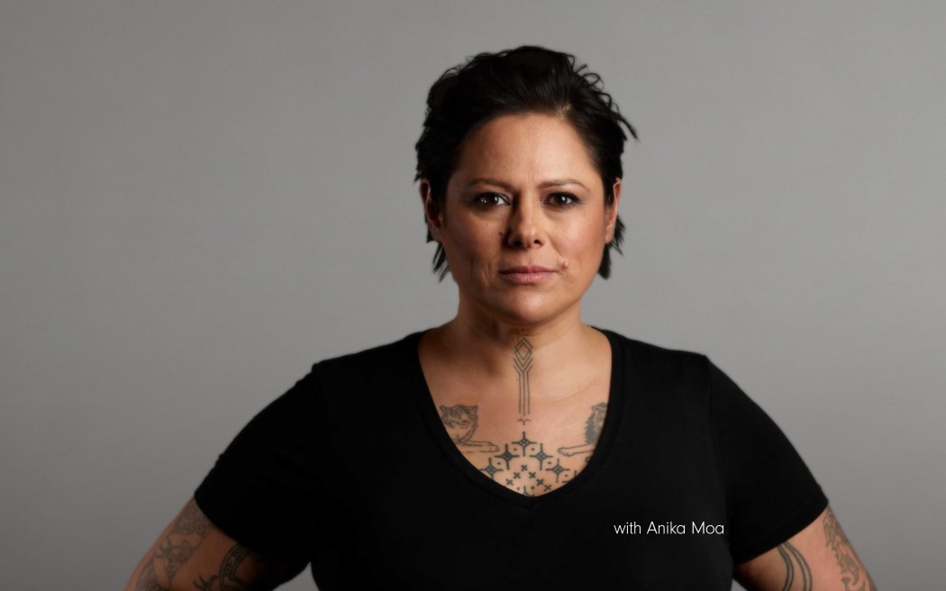 Anika Moa wears a black top with hands on hips, looking straight ahead to camera, on a light grey background.