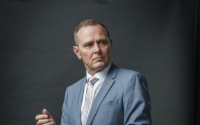 Actor Stephen Lovatt stands dressed in blue suit against a soft grey background.