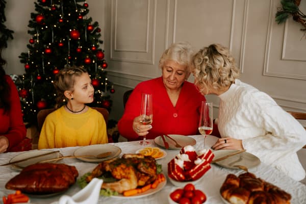 An older women looking down at the wine glass on the dinner table as another woman whispers to her and a child looks at them.