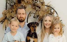 A family portrait. Mum Fallon and Dad Tom sit with their two kids on their knees, and their dog in between them. They all smile at the camera.