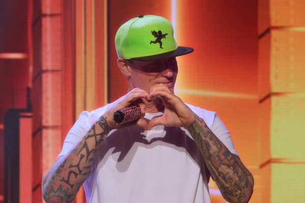 A man with tattooed arms and a lime green hat mades a heart symbol with his fingers.