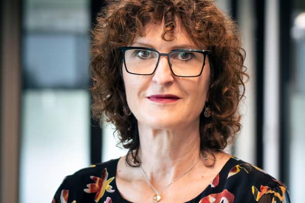 Elisabeth Shaw is a midlife woman weaering dark-rimmed square glasses and curly, shoulder-length brown hair.