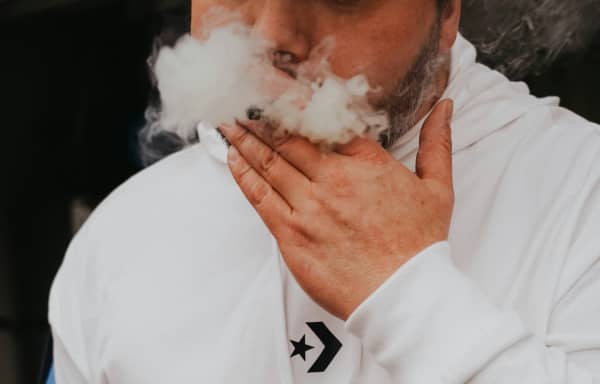 A bearded man in a hoodie inhales smoke.