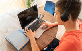 Teenage boy doing homework using computer sitting by desk in room alone.