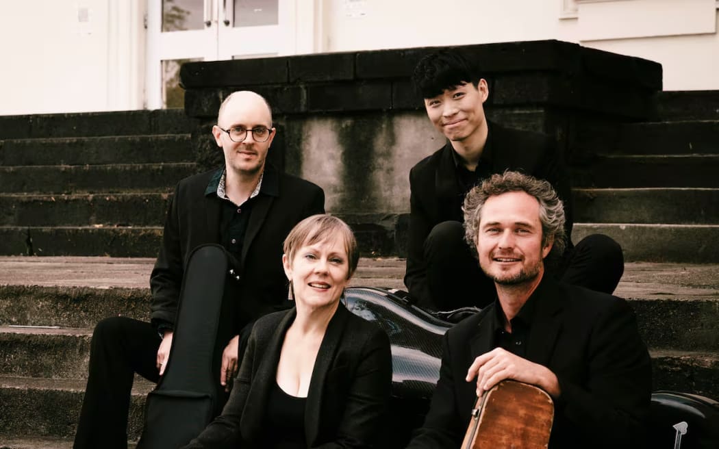 Akarana Piano Quartet members smiling, dressed in black, sit outside on some steps with their instruments in travel cases (but  no piano).