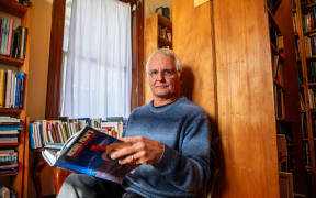Hard to Find Books shop owner Warwick Jordan sits on a small chair in his shop, with a book open in his hands.