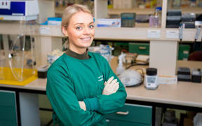 Jemma is smiling and standing with her arms folded in a lab wearing green scrubs and latex gloves