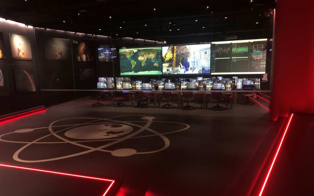 A dark room lit up with neon red tube lighting. A large control room desk sits at the centre, with large screens in front of it, showing live data, a world map, and live feeds from various locations. On the black floor is a large Rocket Lab logo in silver.