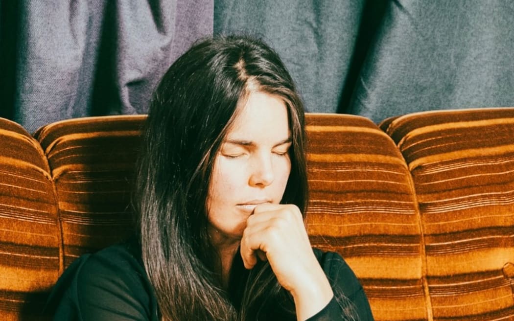Christchurch musician Mel Parsons, a white woman with long dark hair, sits on a brown couch with her eyes closed and a hand up to her mouth