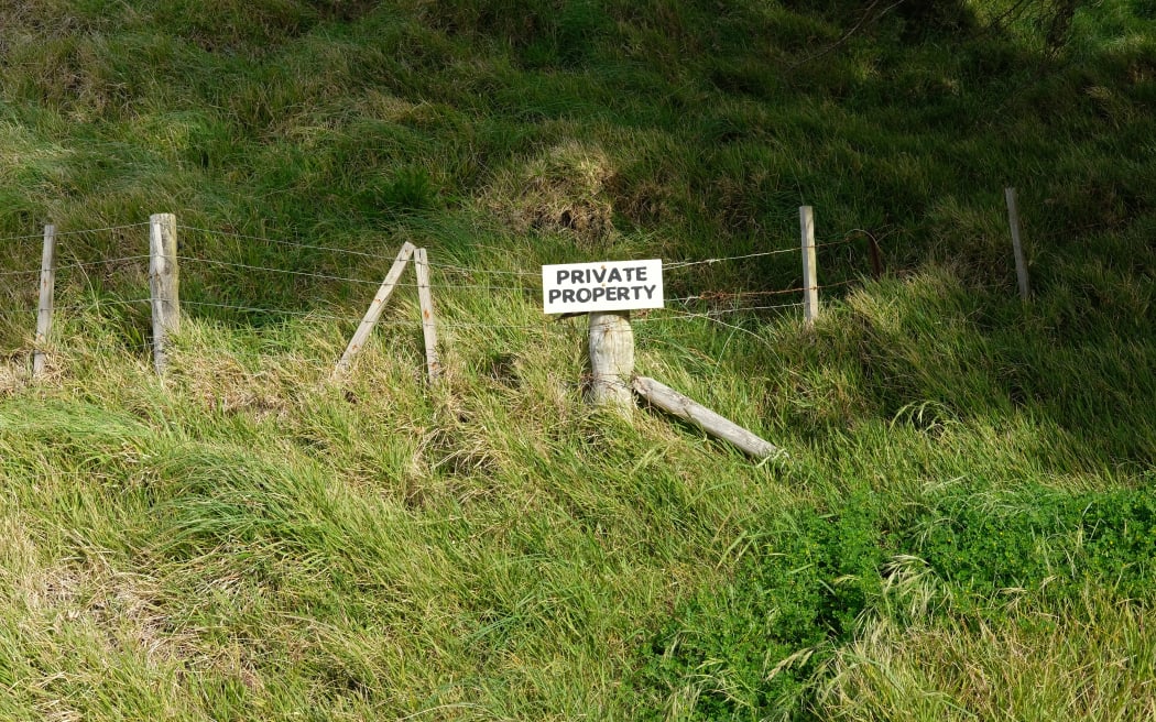 A sign that reads private property is hanging on a barbed wire fence. THe fence cuts across a grassy hill and is likely the boundry of a farm.