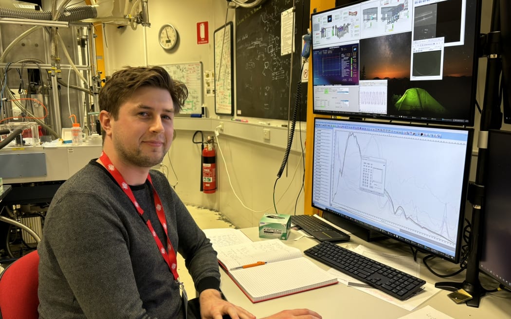A man with a dark sweater and red lanyard sits at a computer monitor. He is smiling at the camera. The monitor displays a graph with a curve on it. He is in a lab with some complicated equipment visible in the background.