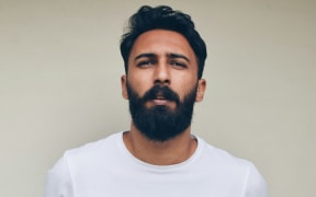 A bearded and brown-skinned and bearded man wears a crisp white t-shirt and stands against a beige wall.