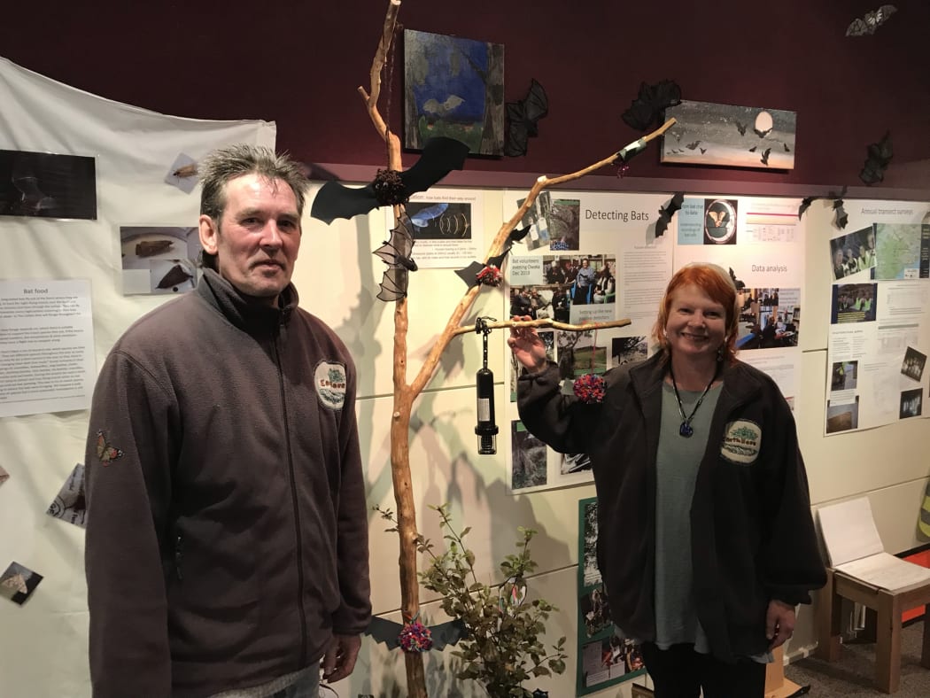 Gordon and Janine Thompson at the Catlins Bats on the Map exhibition in Owaka museum. They stand beside a branch that has bat shapes and a bat detector hanging off it as part of the exhibition display.