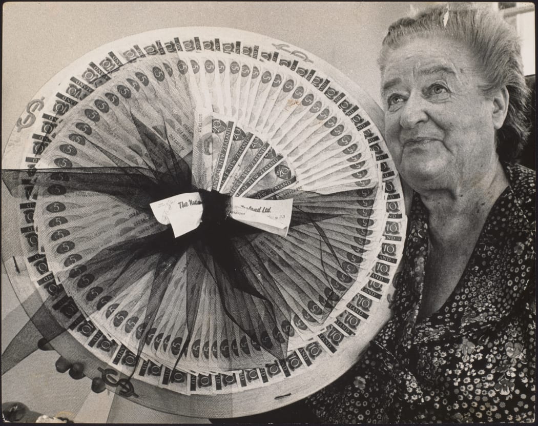 Flora McKenzie with a collection of 5, 10 and 20 dollar notes fanned out in a circle together with a cheque and topped with a bow for payment of a fine. 1973