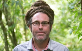 Duncan Kay is standing in front of some trees. it is a closeup head and shoulders shot.