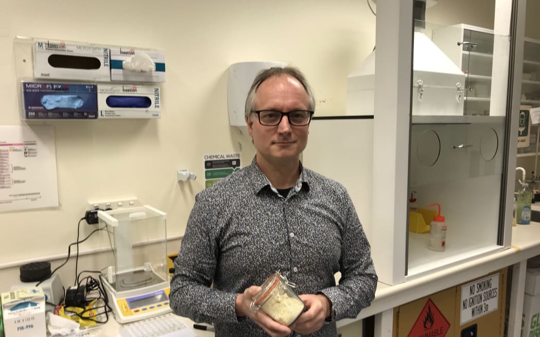 Dr Stefan Hill is standing in front of a lab bench holding a jar with a powdery substance in it.