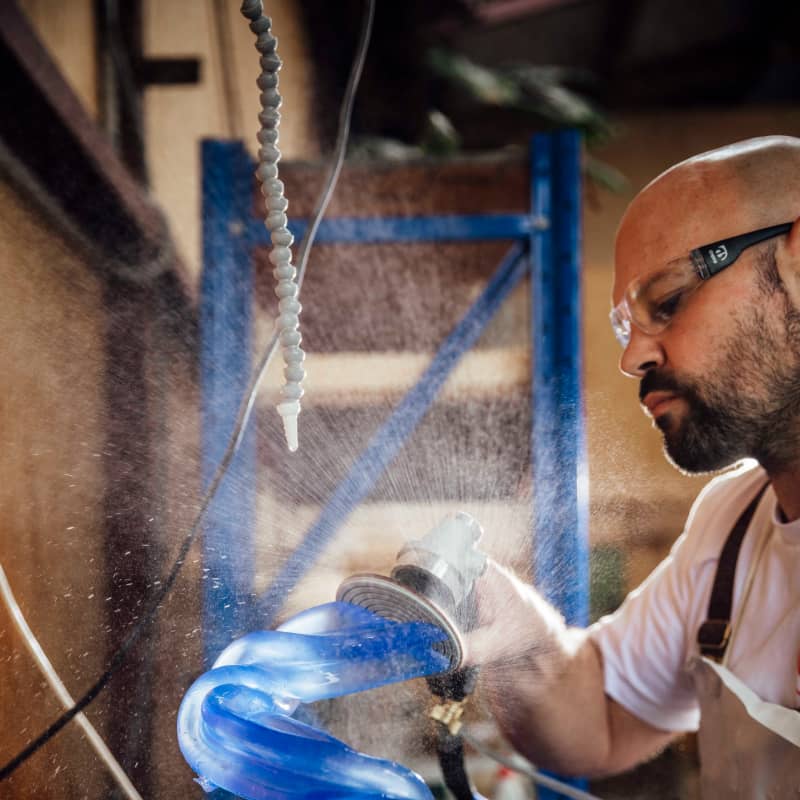A bald, bearded man in safety glasses sculpts a tube of blue glass.