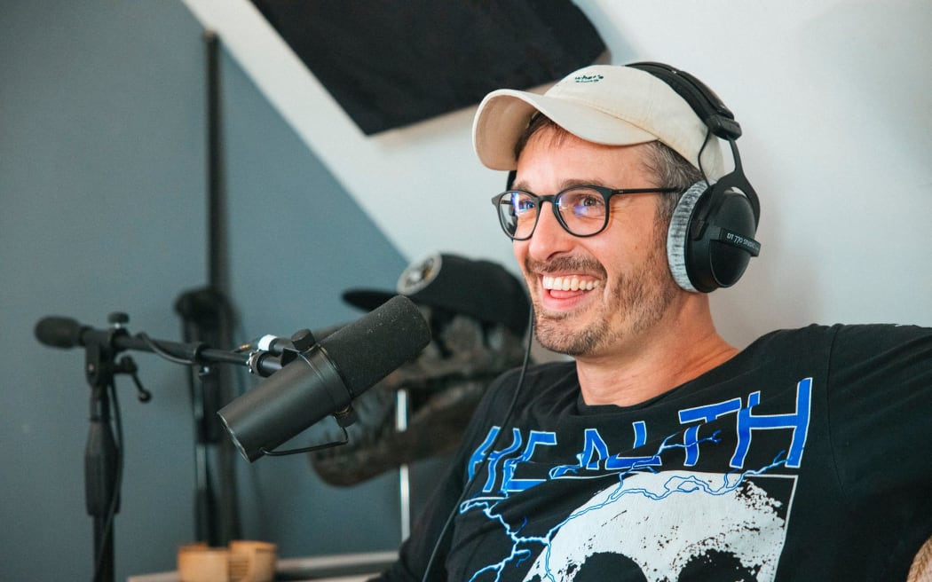 David Farrier wears a hat, glasses and smiles into a microphone