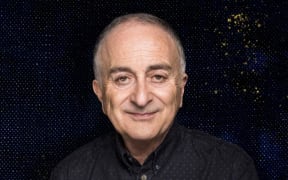 Sir Tony Robinson is a slightly smiling bald man wearing a black shirt with a black background behind him.