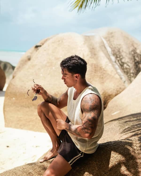 A muscular man with dark hair sits on a rock.