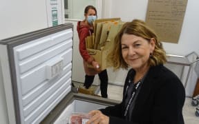 Sophie Gray smiles at the camera. She is packing foodstuffs into a tub freezer at the Good Works Trust food bank. In the background, another worker carries boxes.