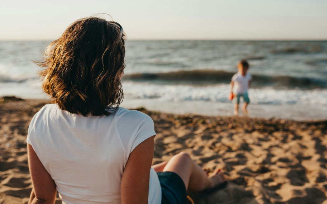 A person in a t-shirt and shorts sits on a beach and a child plays near the water line.