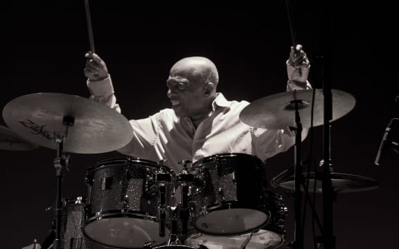 A black and white photo of drummer Roy Haynes in action, lifting his drumsticks in the air and sitting behind a drum set.