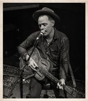 A black and white photo of singer/songwriter C.W Stoneking singing into a microphone and playing a guitar.
