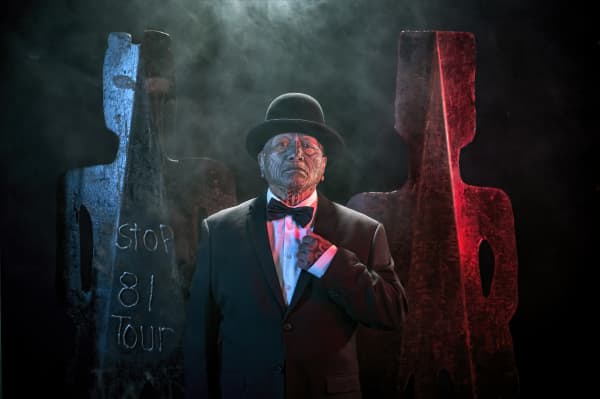 Image from Tame Iti: I Will Not Speak Māori Exhibition.  Artist wearing a suit, bowtie and bowler hat stands in front of metal sculptures in a stylised human form.