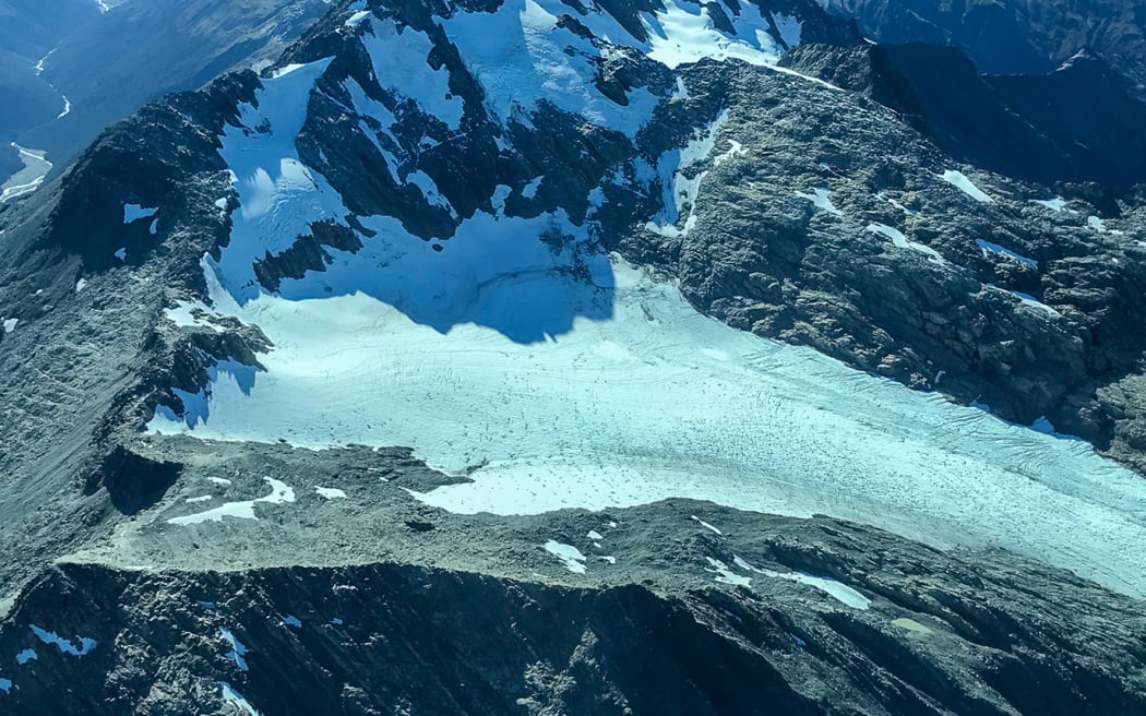 A photo taken from a plane window looking down on a glacier and rocky, snowy mountains, with the underside of the plane wing visible. The sky is blue.