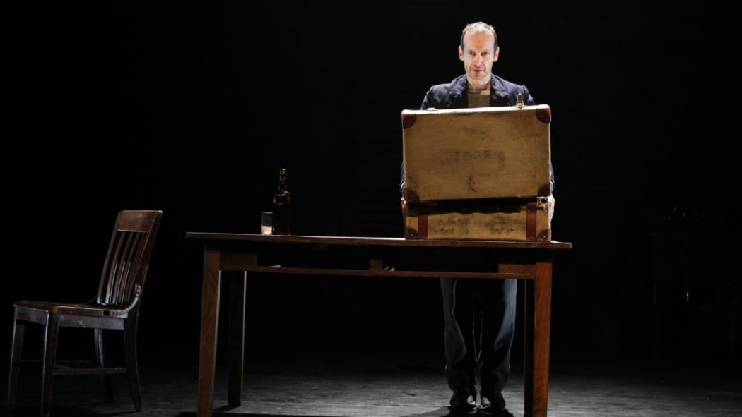 Denis O'Hare stands on a dark stage in front of a suitcase on a table.