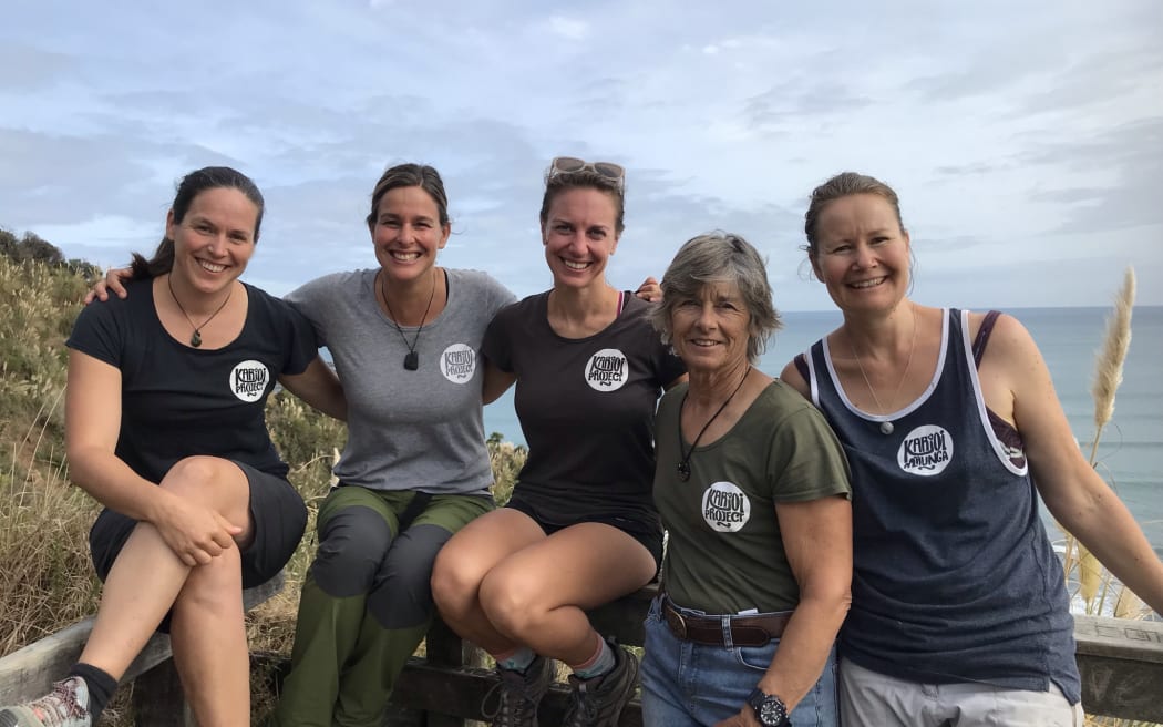 The five members of the team sit or lean against a wooden fence at the look out spot over Ngarunui beach. The ocean is visible behind them.