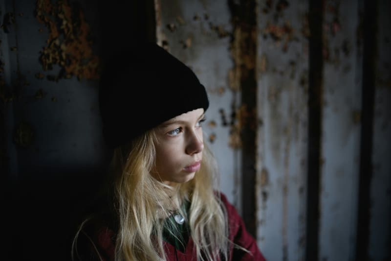 A young blonde girl wearing a black beanie looks out a window.