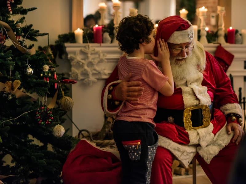 Child whispers into Santa's ear in a room with a fireplace and Christmas tree.