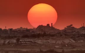 This picture taken from a position on the Israeli border with the Gaza Strip, shows the sun setting behind destroyed buildings in the besieged Palestinian territory on 19 August, 2025.