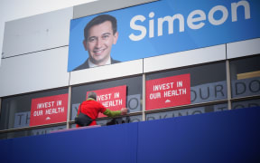 One of the protestors outside Simeon Brown’s electorate office has used a ladder to cover the Health Minister's message in his window with their own placards.