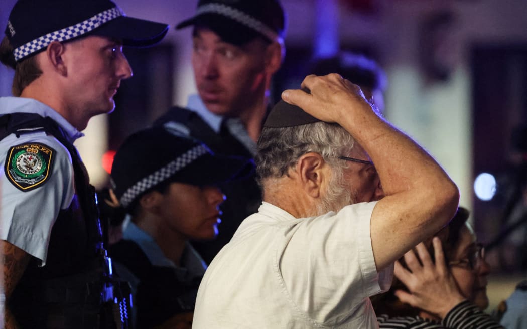 A member of the Jewish community reacts as he walks with police towards the scene of a shooting at Bondi Beach in Sydney on December 14, 2025. Australian police said two people were in custody following reports of multiple gunshots on December 14 at Sydney's famed Bondi Beach, urging the public to take shelter. (Photo by DAVID GRAY / AFP)