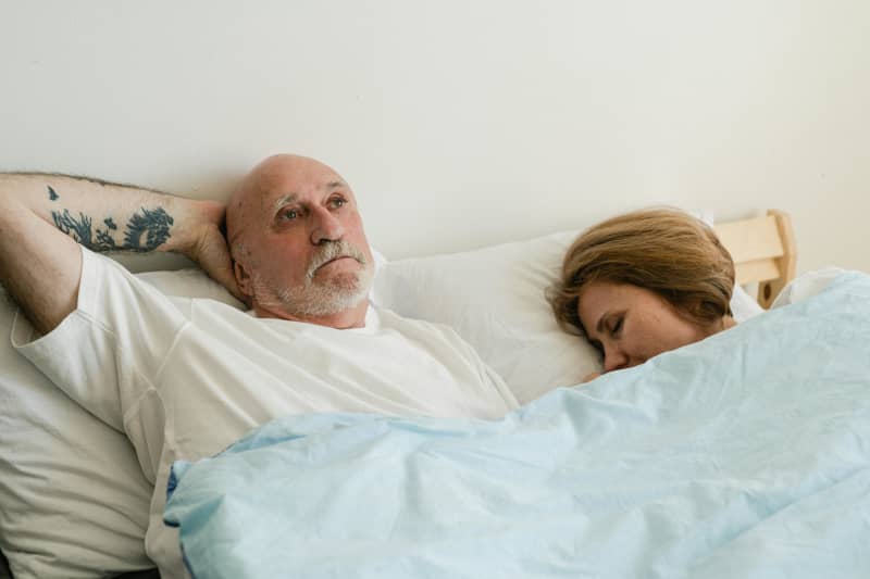 A senior couple are in bed with the woman sleeping and the man awake.