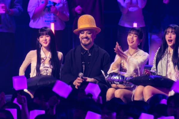 Boy George in a big yellow hat is flanked by three young women.