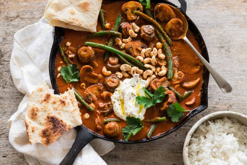 A cast iron frying pan filled with a rich red-brown curry of mushrooms, cashews and green beans, garnished with flatbreads, coriander and yoghurt.