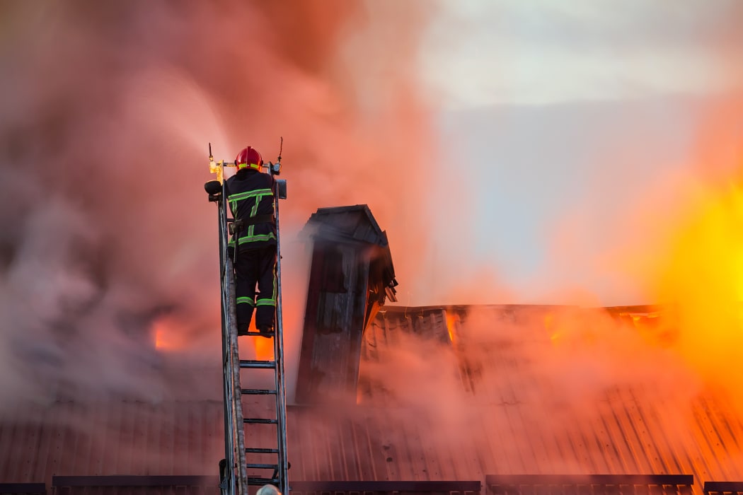 Firefighter or fireman on the ladder extinguishes burning fire flame with smoke on the apartment house roof.