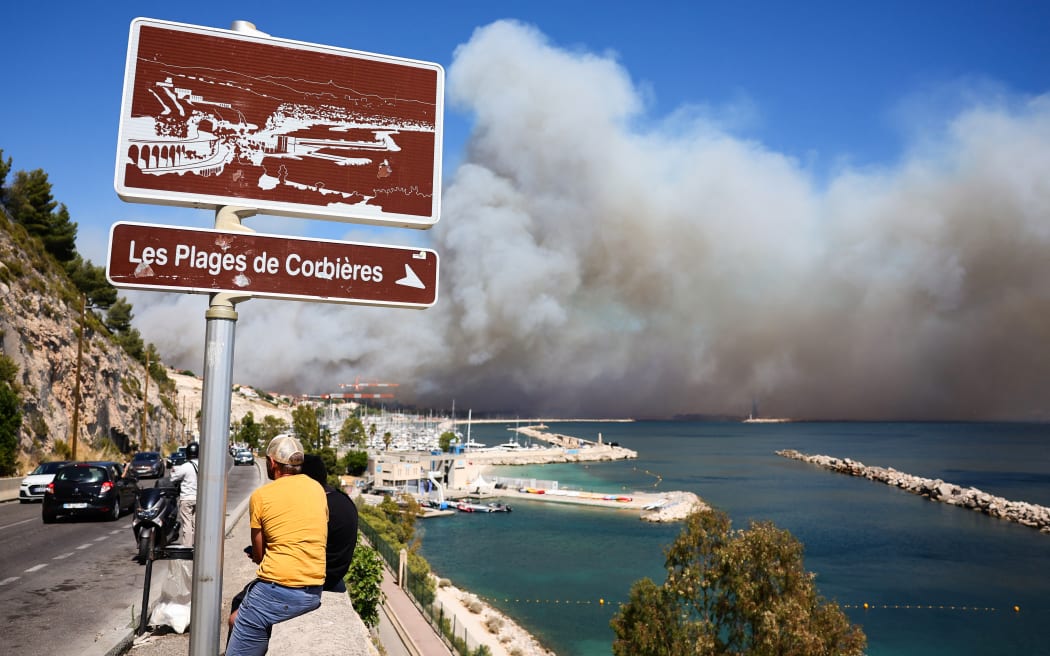 People look on near the Plage des Corbieres, in Marseille, southern France on July 8, 2025, as a smoke from a wildfire rages in the background. The fire that started late on July 8, 2025 morning in Les Pennes-Mirabeau (Bouches-du-Rhone), a town bordering Marseille, has entered France's second-largest city, where some residents were urged to shelter in place, according to a warning message at 4:00 PM from the prefect of Bouches-du-Rhone. (Photo by Clement MAHOUDEAU / AFP)