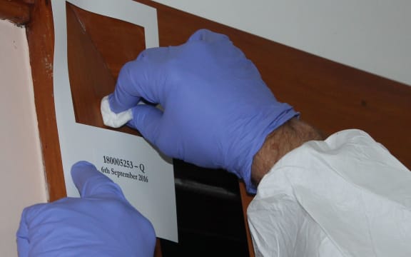 A close up photo of two hands with gloves on, swabbing within a 100 square centimetre stencil stuck to a wall. By swabbing within that area, testing companies get a sample that corresponds to Ministry of Health guidelines for meth.