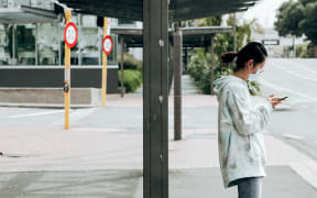 A woman wears a mask as she waits at a bus stop during level 2 in Wellington on15 February 2021
