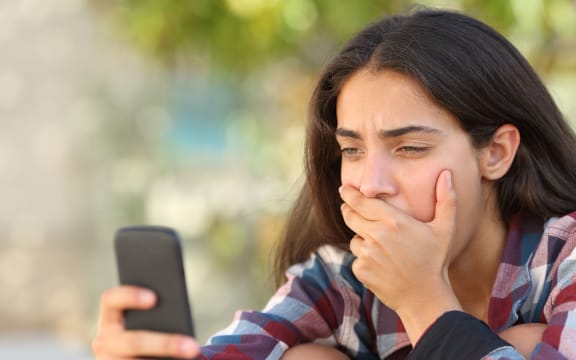 A photo of a worried teenager girl looking at her smart phone in a park with an unfocused background